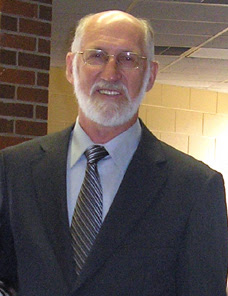 Headshot of Dr. David Voegtlin, who is wearing a dark suit coat, light blue shirt, and striped tie. He is smiling. He has a short white beard and is wearing glasses.